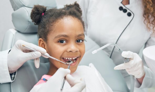 Happy afro american girl sitting in stomatologist chair with open mouth while professional dentist doing regular check up of teeth using dental probe and mirror. Female nurse assisting.